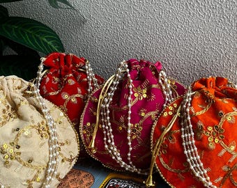 May include: A collection of four velvet drawstring pouches in cream, red, fuchsia, and orange. Each pouch is adorned with gold embroidery and sequins, and features a pearl beaded drawstring. The pouches are displayed on a decorative box.