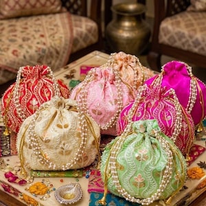 May include: A selection of embroidered drawstring bags in various colours. The bags, in shades of red, pink, green, and cream, feature gold detailing and pearl-like beads. These bags are likely designed for carrying small items.