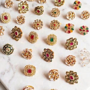May include: A collection of gold-toned rings with various gemstone accents, including red, green, and clear stones. The rings feature intricate floral and geometric designs, displayed on a white marble surface.