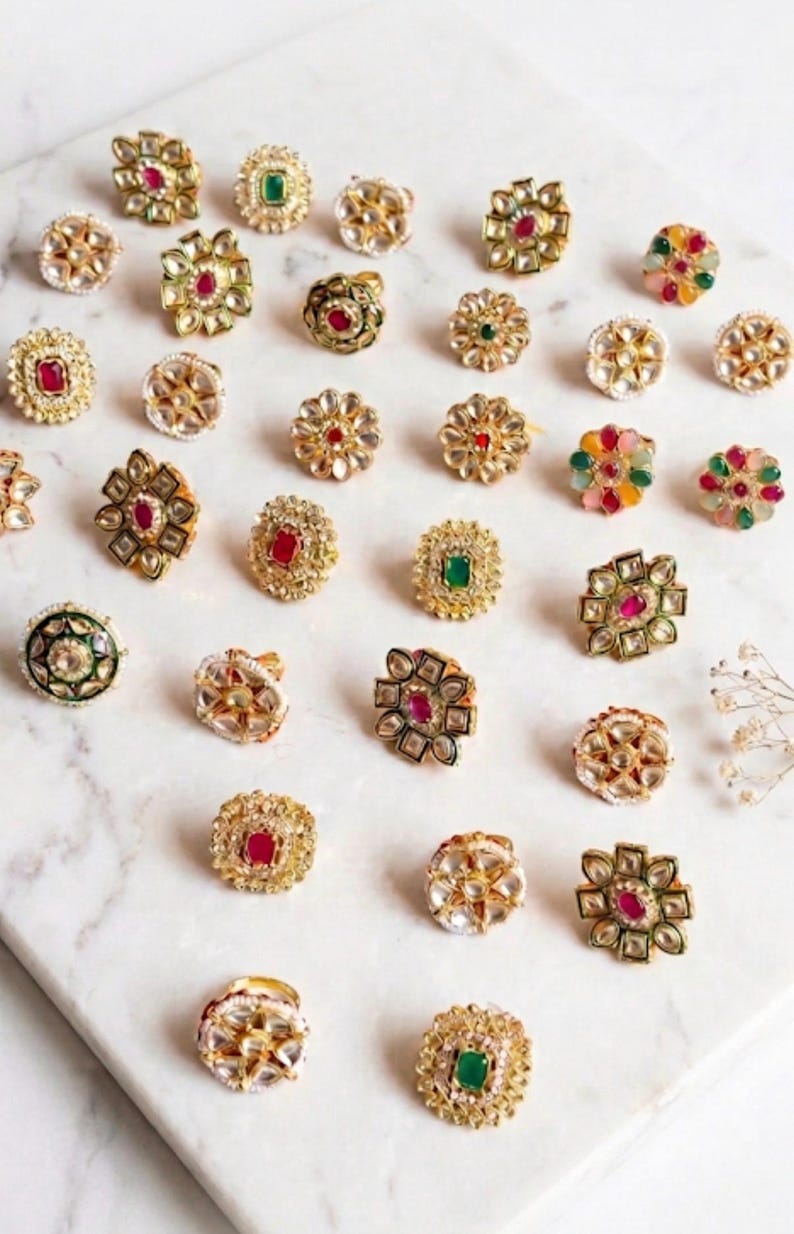 May include: A collection of gold-toned rings with various gemstone accents, including red, green, and clear stones. The rings feature intricate floral and geometric designs, displayed on a white marble surface.