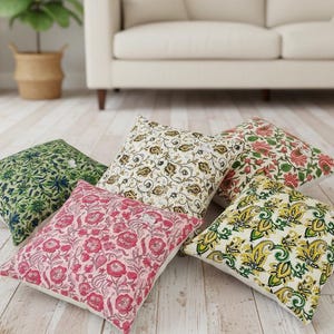 May include: Five square throw pillows with floral and paisley patterns in shades of green, pink, cream, and yellow. The pillows are arranged on a light wood floor, with a neutral-colored sofa in the background.