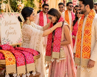 May include: A collection of vibrant red and orange bandhani stoles featuring intricate gold embroidered borders and dangling pearl-like beads. The stoles are displayed on a gold tray next to a sign that reads Welcome Our Guests.
