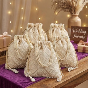 Può includere: Cinque sacchetti avorio con coulisse, decorati con perle e motivi ricamati. Sono disposti su un tavolo di legno con un runner viola. Un cartello recita "Wedding Favours".