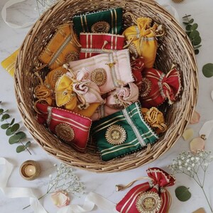 Op de afbeelding: Een rieten mand vol kleurrijke, sierlijke zakjes in geel, roze, rood en groen. Elk zakje heeft gouden versieringen en trekkoorden. Decoratieve kaarsen en bloemstukken omringen de mand, wat een feestelijke uitstraling creëert.