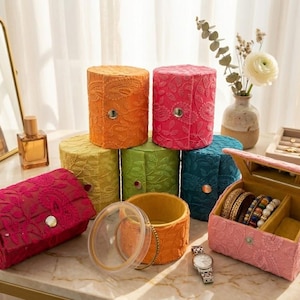May include: A selection of embroidered jewellery boxes in shades of orange, pink, green, and teal. The boxes are cylindrical and rectangular. A pink box is open, displaying bracelets. A watch and perfume bottles are also visible.