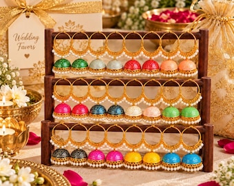 May include: A wooden three-tiered display stand holding twenty-four gold-toned hoop earrings with dangling jhumka-style bell pendants in various colors including green, silver, red, peach, pink, blue, white, black, and yellow.
