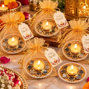 May include: Four gold organza drawstring bags each containing a round mirror diya candle holder with a lit tea light candle. Each bag features a white tag with the text Happy Diwali and a floral illustration. One additional candle holder sits alone.
