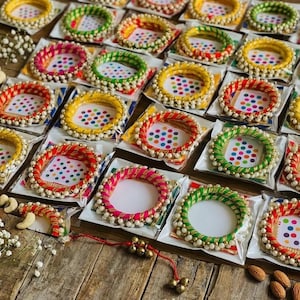 May include: A collection of colorful bangles, each wrapped in vibrant threads of red, pink, green, and yellow. The bangles are adorned with small white beads and presented on a wooden surface, creating a festive display.