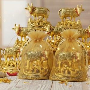 Könnte beinhalten: Goldene Kuhfiguren, einige auf dekorativen Plattformen, andere in durchsichtigen Beuteln. Die Figuren sind aufwendig detailliert, mit glänzendem Finish. Ein kleines Schild zeigt "Handcrafted in India". Das Thema ist religiös oder kulturell.