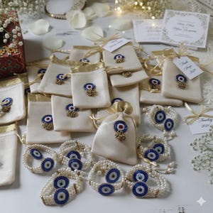 May include: An assortment of ivory-colored drawstring pouches with gold trim and embellishments. Each pouch features a blue and white eye-shaped charm with a red center. Pearl bracelets with matching eye charms are also present.