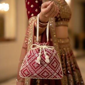 May include: A red embroidered bag with a white floral and geometric pattern. The bag has a drawstring closure and pearl-like beaded handles. The bag is decorated with white tassels and small pearls.