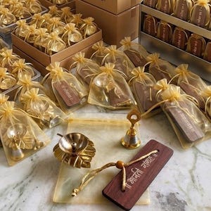 May include: A collection of gold-toned brass diya lamps shaped like leaves, small brass bells, and rectangular wooden plaques with Sanskrit text, each packaged in a sheer gold organza drawstring bag, with some items displayed outside the bags.
