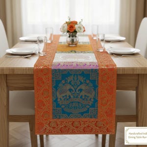 May include: A handcrafted Indian dining table runner with a vibrant design. The runner features a teal central panel with gold elephant motifs, bordered by orange and gold floral patterns. The runner is placed on a wooden table with white plates and a vase of flowers.