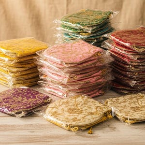 Może przedstawiać: Stosy małych, kwadratowych i okrągłych materiałowych woreczków w różnych kolorach, w tym żółtym, różowym, zielonym, czerwonym, fioletowym i kremowym. Każdy woreczek jest zdobiony złotym haftem i zapakowany w przezroczysty plastik. Woreczki ułożone są na jasnej drewnianej powierzchni.