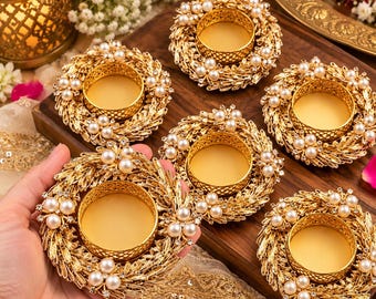 May include: A set of round gold-toned metal candle holders, each featuring a textured wreath-like frame adorned with small white faux pearls and clear crystals. The center of each holder is a smooth, circular gold cup for a tea light candle.