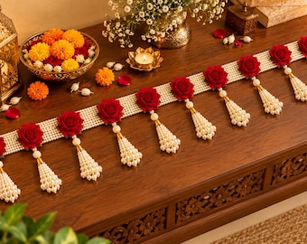 May include: A decorative door hanging featuring a horizontal band of white faux pearls accented with eight vibrant red roses. Each rose is paired with a dangling, cone-shaped tassel crafted from clusters of small white faux pearls.
