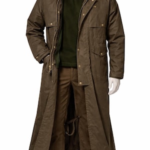 May include: A long, olive-green waxed cotton coat with a corduroy collar, worn over a green sweater and brown pants. The coat has multiple pockets, a zipper closure, and brass-colored buttons. The outfit is displayed on a mannequin.