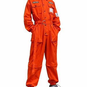 May include: Bright orange jumpsuit with multiple pockets, a belt, and the text "TONSTOUY" on the chest. The jumpsuit has long sleeves and is worn with gray and black sneakers. The overall style is utilitarian.