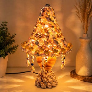 May include: A decorative table lamp made of seashells. The lamp has a conical shade and a base with a textured, coral-like appearance. The shade is adorned with small shells, and the lamp emits a warm, inviting glow. The lamp is approximately 18 inches tall.