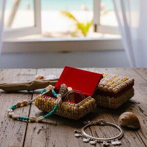 Peut inclure: Une boîte à bijoux en bois avec un intérieur rouge, ouverte pour afficher des bracelets et des bagues. Une boîte fermée assortie est posée à côté. Un collier de coquillages et un bracelet en argent sont également sur la surface en bois. En arrière-plan, une fenêtre avec vue sur la plage.