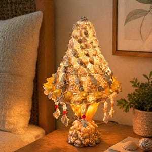 May include: A decorative table lamp made of seashells. The lamp has a conical shade and a base, both covered in various seashells. The lamp is lit, casting a warm glow. Small red and white beads hang from the shade. The lamp sits on a wooden nightstand.