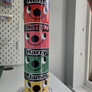 May include: A vertical stack of four ceramic mugs, each featuring a cartoon face and a name. The top mug is red with the name Henry, the second is pink with Hetty, the third is green with Harry, and the bottom is yellow with the name James.