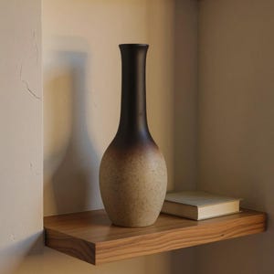 Peut inclure: Un vase décoratif avec un haut brun foncé qui se fond dans une base beige mouchetée. Le vase est exposé sur une étagère en bois avec un livre. Le vase a un long col fin et un corps arrondi.