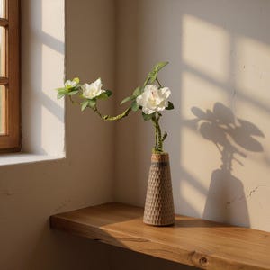 May include: A decorative vase with a textured, brown surface holds a floral arrangement. The arrangement features white flowers and green leaves. The vase sits on a wooden shelf near a window, with sunlight casting shadows on the wall.