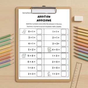 May include: A clipboard with a worksheet titled "ADDITION ADDIZIONE" in black text. The worksheet has addition problems with animal illustrations. Colored pencils, a ruler, and an eraser are on a light wood surface.