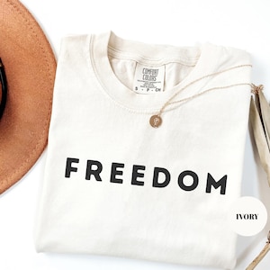 May include: An ivory short-sleeved t-shirt with the word FREEDOM printed in bold black capital letters across the chest. The shirt features a Comfort Colors brand label at the inner neckline and is paired with a delicate gold-toned pendant necklace.