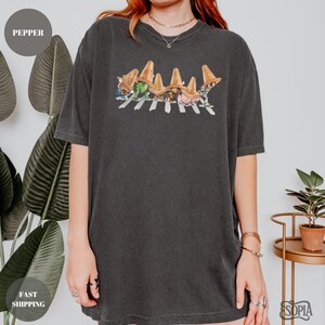 May include: Dark gray t-shirt featuring a colorful graphic of cartoon characters crossing a street, with brown hats. The shirt has a crew neck and short sleeves. The word "Pepper" is in a circle in the upper left corner.