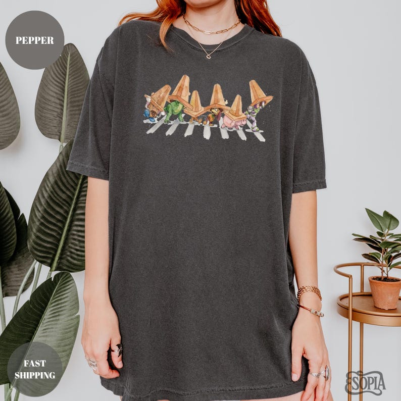 May include: Dark gray t-shirt featuring a colorful graphic of cartoon characters crossing a street, with brown hats. The shirt has a crew neck and short sleeves. The word "Pepper" is in a circle in the upper left corner.