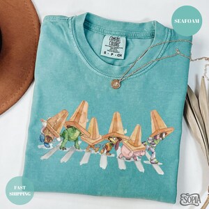 May include: Seafoam green t-shirt with a graphic of cartoon characters crossing a street, each wearing a traffic cone hat. The shirt has a Comfort Colors tag and a gold necklace. The shirt is folded on a surface.