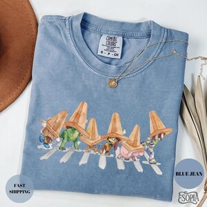 May include: Blue jean-colored t-shirt featuring a whimsical illustration of cartoon characters crossing a street, each wearing a traffic cone. The shirt has a crew neck and short sleeves. The text "Comfort Colors" is visible on the tag.