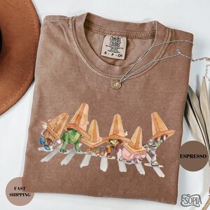 May include: Espresso-colored Comfort Colors t-shirt with a graphic of cartoon characters crossing a crosswalk, each wearing a traffic cone. The shirt has a crew neck and short sleeves. A silver necklace with a pendant is draped on the shirt.