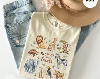 May include: Ivory-colored t-shirt featuring a colorful illustration of various African animals, including a lion, cheetah, and zebra. The text "Respect the locals" is printed below the animals. The shirt is paired with light blue jeans and a beige hat.