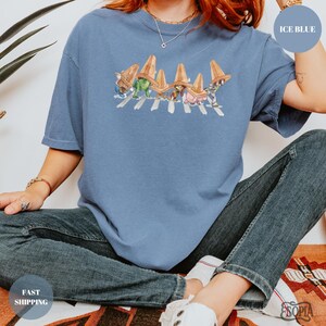 May include: Ice blue t-shirt featuring a graphic of cartoon characters crossing a crosswalk, wearing sombreros. The shirt is a light blue color. The text "ICE BLUE" is in a circle on the upper right. The text "FAST SHIPPING" is in a circle on the lower left.