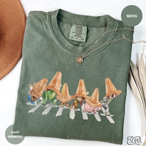 May include: Moss green t-shirt with a graphic of cartoon characters wearing traffic cones and crossing a crosswalk. The shirt has a Comfort Colors tag and a small necklace. The shirt is folded on a white surface.
