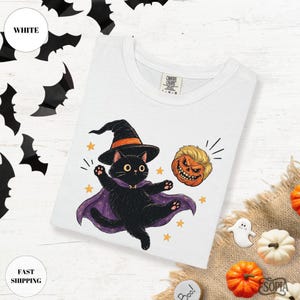 May include: A white t-shirt featuring a graphic of a black cat wearing a witch hat and a purple cape, floating next to a carved pumpkin with blonde hair. The shirt has a Comfort Colors label at the neckline and is surrounded by bat cutouts.
