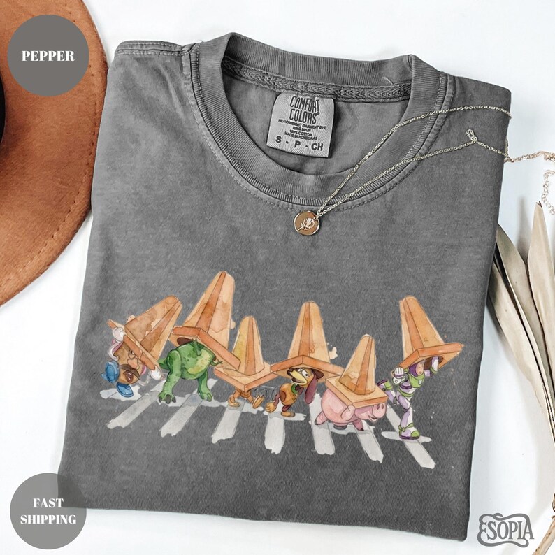 May include: Gray Comfort Colors t-shirt featuring a whimsical illustration of cartoon characters crossing a street, each wearing a traffic cone hat. A silver necklace with a pendant is draped on the shirt.