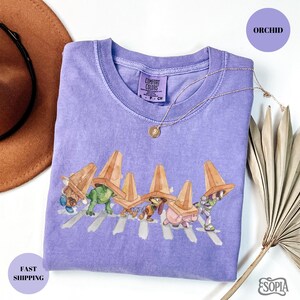 May include: Orchid-colored t-shirt featuring a whimsical illustration of cartoon characters crossing a street, each wearing a traffic cone hat. The shirt has a crew neck and short sleeves. A gold necklace with a pendant is draped on the shirt.