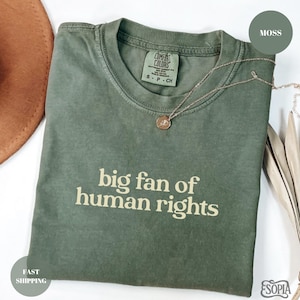 Peut inclure: Un t-shirt vert mousse plié avec l'inscription "big fan of human rights" en beige clair. Le t-shirt porte une petite étiquette au niveau de l'encolure. Un collier doré avec un pendentif est posé sur le t-shirt.
