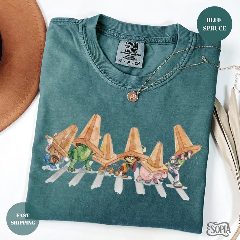 May include: Blue spruce Comfort Colors t-shirt featuring a graphic of cartoon characters crossing a crosswalk, each wearing a traffic cone hat. A gold necklace with a pendant is draped across the shirt. The shirt is folded.