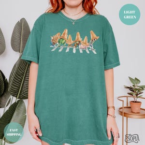 May include: A teal green t-shirt featuring a whimsical graphic of cartoon characters wearing traffic cones and crossing a crosswalk. The shirt has a relaxed fit and short sleeves. The text "LIGHT GREEN" is in a circle on the upper right.