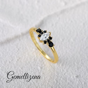 Può includere: Un anello dorato con una pietra centrale trasparente a forma di marquise, circondata da gruppi di piccole pietre nere. L'anello ha una fascia sottile e lucida. La parola "Gemellizena" è scritta in corsivo in basso.