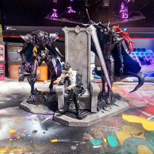 May include: A detailed fantasy figurine set featuring a central figure seated on a throne, flanked by two armored figures. The figures are dark with purple accents, and the throne is gray. The background includes a computer monitor and a colorful surface.