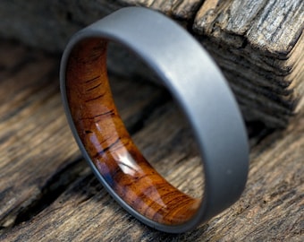May include: A grey titanium ring with a brown wood inlay. The ring is on a wooden surface.