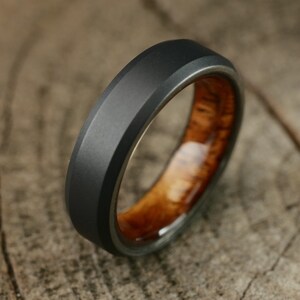 Puede incluir: Un anillo negro y marrón con un acabado de metal cepillado y una incrustación de madera. El anillo está sobre una superficie de madera.