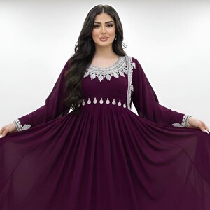May include: A long, flowing, plum-colored dress with long sleeves. The dress features intricate silver embroidery around the neckline, cuffs, and bodice. The dress has a gathered waist and a full skirt, creating an elegant silhouette.