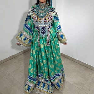 Peut inclure: Longue robe fluide de couleur sarcelle avec des accents dorés et des broderies complexes. La robe présente un col montant orné de perles turquoise et d'ornements colorés. Les manches sont longues et évasées, avec des motifs détaillés.